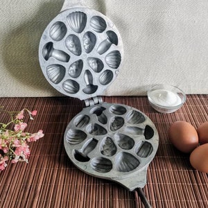 May include: A silver metal mold with a hinged lid, featuring multiple heart-shaped cavities. The mold is open, revealing the cavities. Three brown eggs and a small bowl of white powder are also visible.