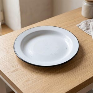 Puede incluir: Un plato redondo de esmalte blanco con un borde fino de color negro. El plato tiene una superficie lisa y brillante con un borde ancho y plano. El diseño es sencillo y circular, con un centro blanco que contrasta con el borde oscuro.