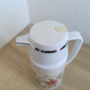 May include: A white insulated carafe with a floral design featuring red and yellow flowers. The carafe has a white lid with a light yellow lever, a silver spout, and a white handle. The carafe is designed to keep beverages hot or cold.