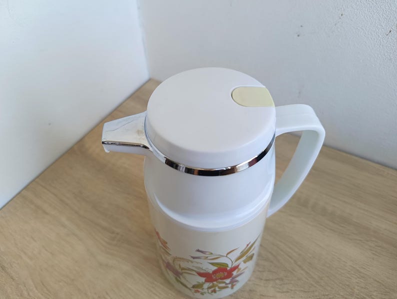 May include: A white insulated carafe with a floral design featuring red and yellow flowers. The carafe has a white lid with a light yellow lever, a silver spout, and a white handle. The carafe is designed to keep beverages hot or cold.