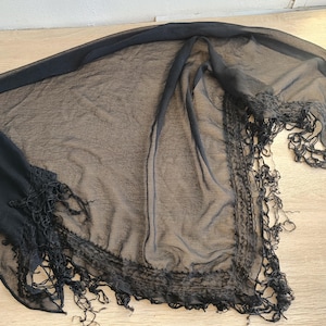 May include: A sheer black shawl with a delicate, fringed edge. The lightweight fabric drapes across a light-colored surface, showcasing the intricate details of the fringe and the subtle texture of the material. This accessory adds an elegant touch.