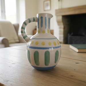 May include: A white ceramic pitcher with a rounded body and a curved handle. The pitcher is decorated with blue, yellow, and green painted designs. The pitcher is sitting on a wooden table.