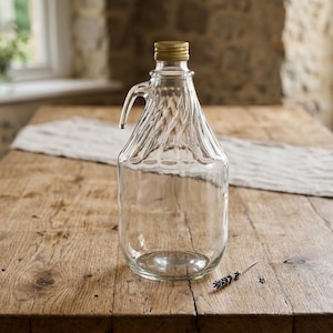 May include: A clear glass jug with a gold-colored metal cap and a handle. The jug has a textured, ridged design on the upper portion. It sits on a wooden table with a light-colored runner. A sprig of lavender lies on the table.
