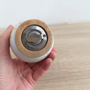Op de afbeelding: Een witte keramische zout- of pepermolen met een houten basis en een roestvrijstalen maalmechanisme. De molen heeft een afgeronde vorm en is versierd met kleine bloemmotieven in geel en blauw.