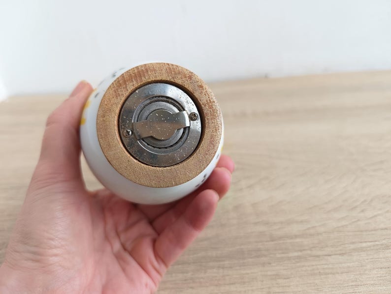 Op de afbeelding: Een witte keramische zout- of pepermolen met een houten basis en een roestvrijstalen maalmechanisme. De molen heeft een afgeronde vorm en is versierd met kleine bloemmotieven in geel en blauw.