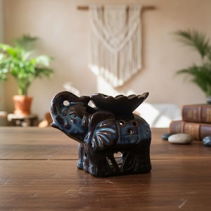 Könnte beinhalten: Ein Elefanten-Öldiffusor aus Keramik in Blau- und Brauntönen steht auf einer Holzoberfläche. Der Elefant hat eine dekorative Schale und ausgeschnittene Designs. Im Hintergrund sind Pflanzen und Bücher zu sehen.