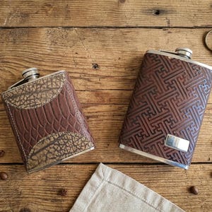 Può includere: Due fiaschette in pelle marrone con dettagli argentati su una superficie di legno. Una ha un motivo, l'altra una texture simile a quella di un rettile. Chicchi di caffè e chiavi sparsi.