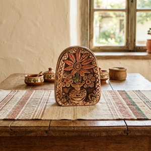 May include: A carved, brown, decorative plaque with a floral design, featuring a large flower and vase. The plaque is on a wooden table with other ceramic items and a striped runner. A window is in the background.