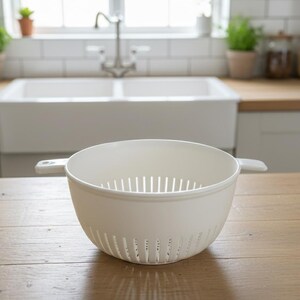 May include: A white plastic colander with a round shape and two handles. The colander has vertical slots for draining and is sitting on a wooden surface. The background includes a kitchen sink and window.