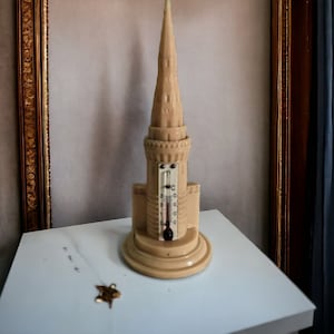 Peut inclure: Un thermomètre en céramique blanche en forme de tour avec un toit pointu. Le thermomètre a une échelle noire et blanche et un pointeur noir. Le thermomètre est posé sur une table blanche.