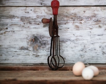 Può includere: Un frullatore per uova vintage a manovella con manico rosso e tre fruste in metallo. Tre uova sono in primo piano su una superficie di legno.