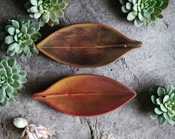 May include: Two leaf-shaped wooden trays in shades of brown and red-orange, each with a small hole at the tip. The trays are surrounded by green succulent plants on a grey, textured surface. A natural, earthy aesthetic.