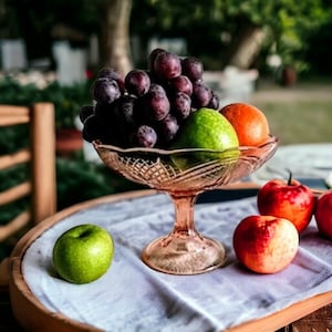 May include: A glass bowl filled with grapes and green and orange fruit. The bowl is sitting on a light brown table with a white tablecloth. There are three red apples on the table.