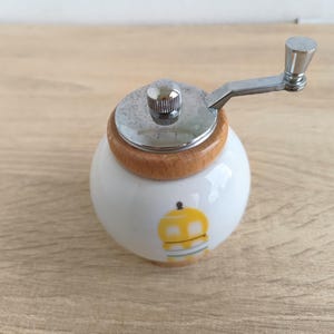 A small, round vintage-style pepper mill with a white porcelain body and a light wooden top. The mill features a subtle yellow geometric graphic on its side and has an exposed metal grinding mechanism at the top. The image is a close-up, showcasing the mill's design and internal structure, resting on a rustic farmhouse wooden table.
