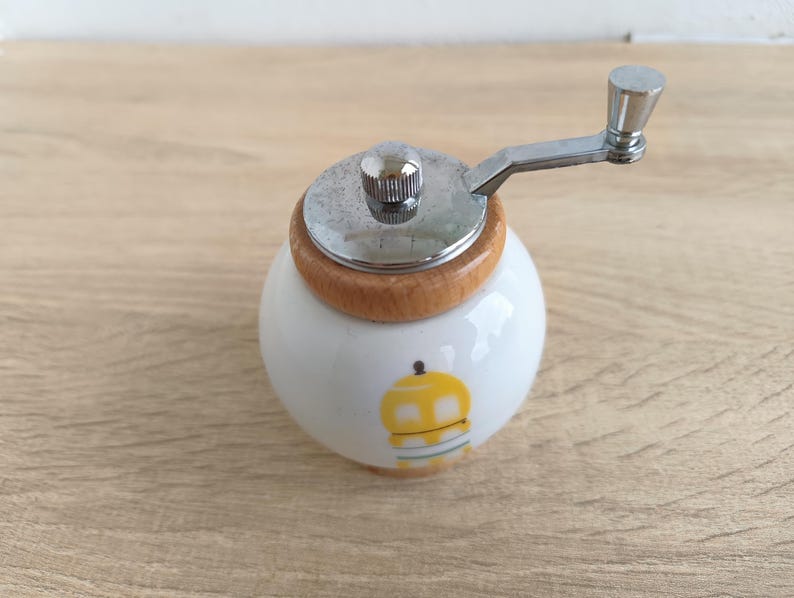 A small, round vintage-style pepper mill with a white porcelain body and a light wooden top. The mill features a subtle yellow geometric graphic on its side and has an exposed metal grinding mechanism at the top. The image is a close-up, showcasing the mill's design and internal structure, resting on a rustic farmhouse wooden table.
