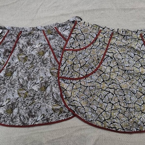 以下が含まれることがあります： 赤色の縁取りが付いた、白と茶色の模様の丸いエプロンが2つ。左側のエプロンには、「LIDA」と書かれた白いタグが付いています。