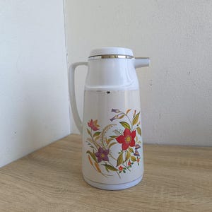 Vintage cream-colored thermos jug with a vibrant retro floral print featuring a red flower, purple blossoms, and wheat. The insulated carafe has a white plastic handle, lid, and pouring spout, accented by a shiny chrome metal trim. Inside, a reflective silver glass vacuum flask is visible. The white plastic screw-top lid has a small cream push button on top. The base is white with a large black plastic central screw underneath holding the flask secure