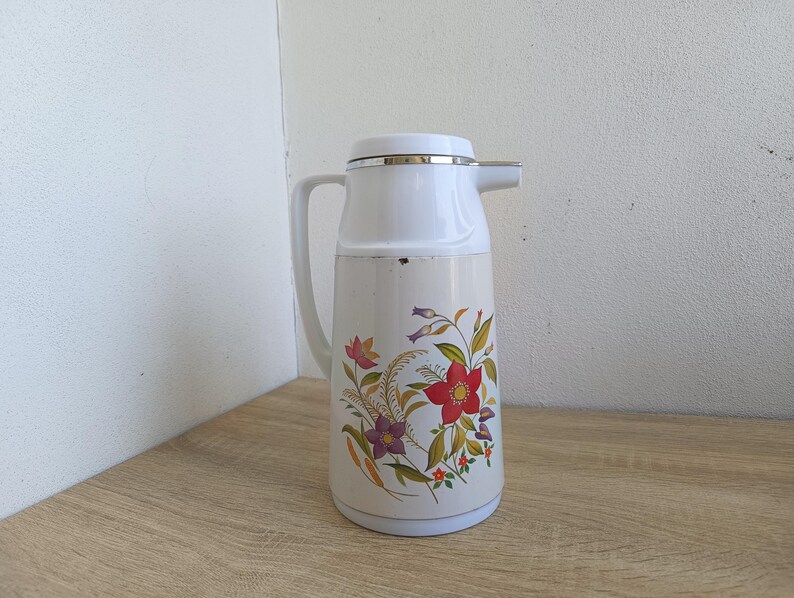 Vintage cream-colored thermos jug with a vibrant retro floral print featuring a red flower, purple blossoms, and wheat. The insulated carafe has a white plastic handle, lid, and pouring spout, accented by a shiny chrome metal trim. Inside, a reflective silver glass vacuum flask is visible. The white plastic screw-top lid has a small cream push button on top. The base is white with a large black plastic central screw underneath holding the flask secure