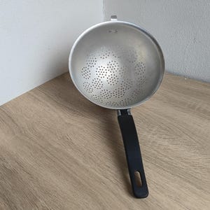 May include: A silver metal colander with a black handle. The colander has a bowl shape with many small holes for draining liquids. The handle is attached to the side of the bowl and has a loop at the end for hanging.