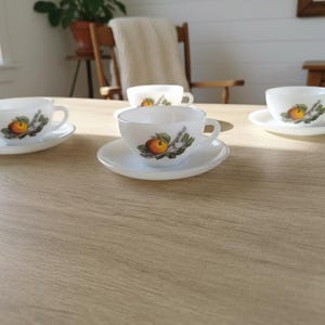 May include: Four white teacups and saucers are arranged on a light brown wooden table. Each cup features a colorful illustration of a peach with green leaves. The scene is set in a well-lit room.
