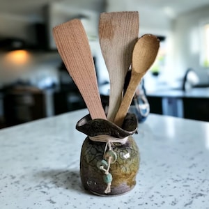 Puede incluir: Un jarrón de cerámica con un esmalte craquelado en tonos marrones y verdes, que contiene tres utensilios de cocina de madera: una cuchara, una espátula y una cuchara de madera.