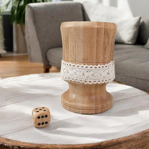 May include: A wooden cup with a lace trim sits on a white wooden table with a black dice beside it.