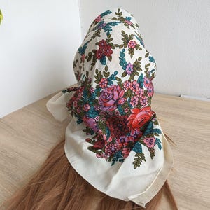Può includere: Un foulard color crema con un motivo floreale con rose rosa, rosse e viola, insieme a foglie verdi. Il foulard è drappeggiato su capelli castani, creando un'esposizione visivamente accattivante.