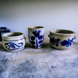 Puede incluir: Tres macetas de cerámica con diseños florales azules y blancos. Las macetas son de diferentes tamaños y formas, y todas están sobre una superficie gris claro.