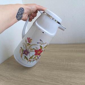 Vintage cream-colored thermos jug with a vibrant retro floral print featuring a red flower, purple blossoms, and wheat. The insulated carafe has a white plastic handle, lid, and pouring spout, accented by a shiny chrome metal trim. Inside, a reflective silver glass vacuum flask is visible. The white plastic screw-top lid has a small cream push button on top. The base is white with a large black plastic central screw underneath holding the flask secure