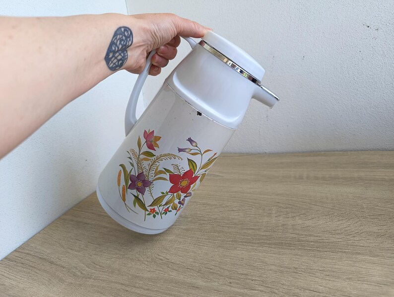 Vintage cream-colored thermos jug with a vibrant retro floral print featuring a red flower, purple blossoms, and wheat. The insulated carafe has a white plastic handle, lid, and pouring spout, accented by a shiny chrome metal trim. Inside, a reflective silver glass vacuum flask is visible. The white plastic screw-top lid has a small cream push button on top. The base is white with a large black plastic central screw underneath holding the flask secure