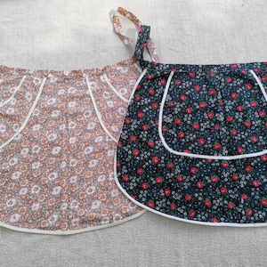 以下が含まれることがあります： 白いトリムが付いた、花柄のヴィンテージエプロン2枚。1枚は白の花がついた茶色、もう1枚は赤と白の花がついた黒です。