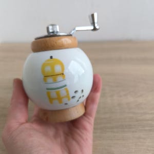 A small, round vintage-style pepper mill with a white porcelain body and a light wooden top. The mill features a subtle yellow geometric graphic on its side and has an exposed metal grinding mechanism at the top. The image is a close-up, showcasing the mill's design and internal structure, resting on a rustic farmhouse wooden table.
