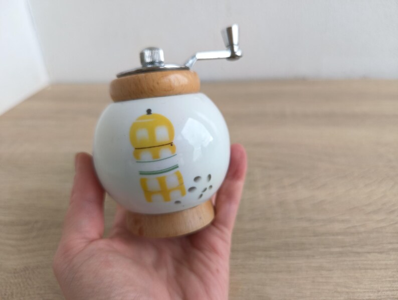 A small, round vintage-style pepper mill with a white porcelain body and a light wooden top. The mill features a subtle yellow geometric graphic on its side and has an exposed metal grinding mechanism at the top. The image is a close-up, showcasing the mill's design and internal structure, resting on a rustic farmhouse wooden table.