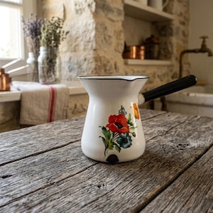 Peut inclure: Cafetière en émail blanc avec une poignée noire et un motif floral. Le pot présente une fleur de pavot rouge, des fleurs orange et bleues. Il est posé sur une table en bois vieilli dans une cuisine rustique.