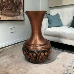 Puede incluir: Un jarrón alto de color cobre, con una base ancha y una parte superior acampanada. La base presenta un diseño floral en relieve y detalles en forma de cuerda. Un elemento decorativo para el hogar, adecuado para salones o entradas.