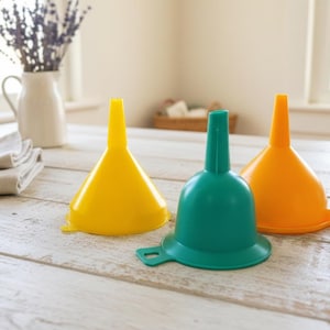 Puede incluir: Tres embudos de plástico en amarillo, verde azulado y naranja se muestran sobre una superficie de madera clara. Los embudos tienen forma de cono con una boquilla estrecha. El fondo incluye una jarra blanca con lavanda y una cesta.