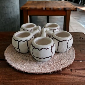 Puede incluir: Cinco pequeñas tazas de cerámica blanca con ribetes marrones, dispuestas en círculo sobre una estera de paja tejida. Las tazas tienen un diseño único con líneas marrones que forman un patrón.