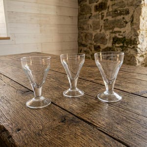Puede incluir: Tres copas transparentes con un diseño acampanado único están dispuestas sobre una mesa de madera rústica. Las copas tienen forma de cuenco cónico y una base sólida. La superficie de la mesa tiene un aspecto envejecido, lo que añade a la estética vintage.