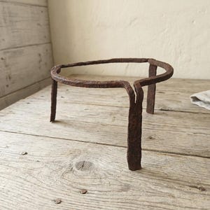 Può includere: Un supporto per piante rustico in ferro battuto, con una parte superiore circolare e tre gambe. Il supporto è di colore marrone scuro invecchiato, suggerendo che sia vintage. Si trova su una superficie di legno usurata.