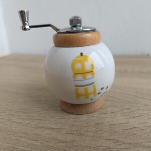 A small, round vintage-style pepper mill with a white porcelain body and a light wooden top. The mill features a subtle yellow geometric graphic on its side and has an exposed metal grinding mechanism at the top. The image is a close-up, showcasing the mill's design and internal structure, resting on a rustic farmhouse wooden table.