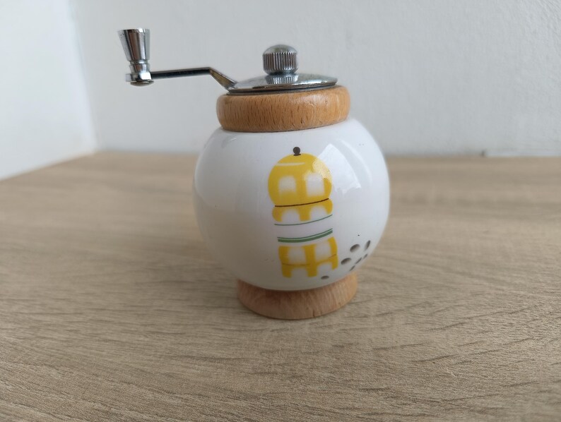 A small, round vintage-style pepper mill with a white porcelain body and a light wooden top. The mill features a subtle yellow geometric graphic on its side and has an exposed metal grinding mechanism at the top. The image is a close-up, showcasing the mill's design and internal structure, resting on a rustic farmhouse wooden table.