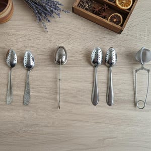 Peut inclure: Vue aérienne de différents infuseurs et passoires à thé. On distingue plusieurs cuillères à thé argentées perforées, un infuseur à long manche et une passoire articulée. De la lavande séchée, des épices et des tasses en bois sont également visibles.