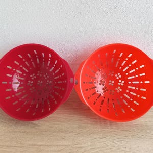 Peut inclure: Deux passoires rondes en plastique rouge et orange avec des poignées. Chaque passoire comporte de nombreux petits trous pour l'égouttage. Les passoires sont sur une surface en bois.