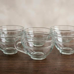 May include: A set of five clear glass teacups arranged in a group. Each cup features a smooth, transparent finish with two horizontal embossed rings circling the exterior body and a curved handle attached to the side of the circular bowl.