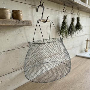 Op de afbeelding: Een draadgaasmand in vintage-stijl met een houten handvat hangt aan een metalen haak. De mand heeft een afgeronde vorm en hangt tegen een achtergrond van een rustieke, wit geschilderde houten muur. Gedroogde kruiden hangen in de buurt.