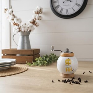 A small, round vintage-style pepper mill with a white porcelain body and a light wooden top. The mill features a subtle yellow geometric graphic on its side and has an exposed metal grinding mechanism at the top. The image is a close-up, showcasing the mill's design and internal structure, resting on a rustic farmhouse wooden table.