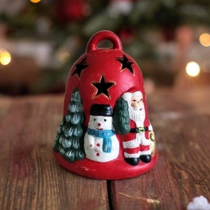 May include: A red ceramic Christmas bell with star-shaped cutouts. The bell features a snowman, a Christmas tree, and Santa Claus. The bell has a loop at the top for hanging and is placed on a wooden surface.