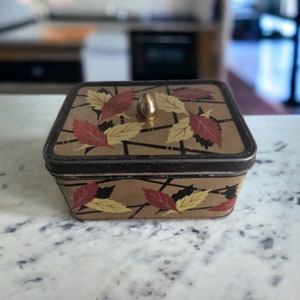 Op de afbeelding: Een vintage rechthoekige blikken doos met een geometrisch ontwerp in goud en bruin, met gestileerde bladeren in tinten rood, geel en zwart. De doos heeft een gouden metalen knop aan de bovenkant.