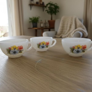 Puede incluir: Tres tazas de té blancas con asas, cada una adornada con un diseño floral con flores amarillas, rojas y azules. Las tazas están dispuestas sobre una mesa de madera clara, lo que sugiere una estética vintage o cottagecore.