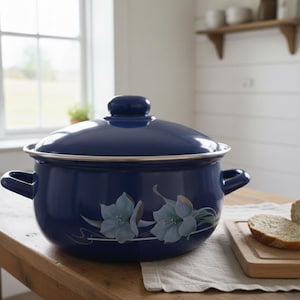 以下が含まれることがあります： 蓋と2つのハンドルが付いた、ネイビーブルーのエナメル鍋。鍋には、水色と白の花柄のデザインが施されています。木製のテーブルの上に置かれ、パンが入ったまな板の隣にあります。